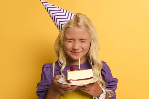 Smiling kid holding plate with birthday cake and making wish on yellow background — Stock Photo
