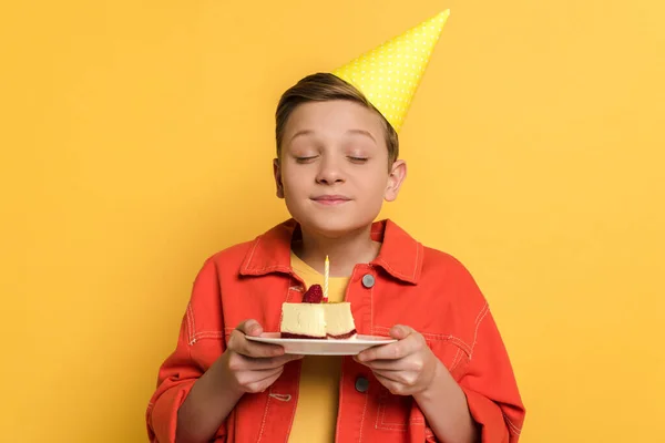 Bambino sorridente a occhi chiusi che esprime desiderio e tiene il piatto con torta di compleanno su sfondo giallo — Foto stock