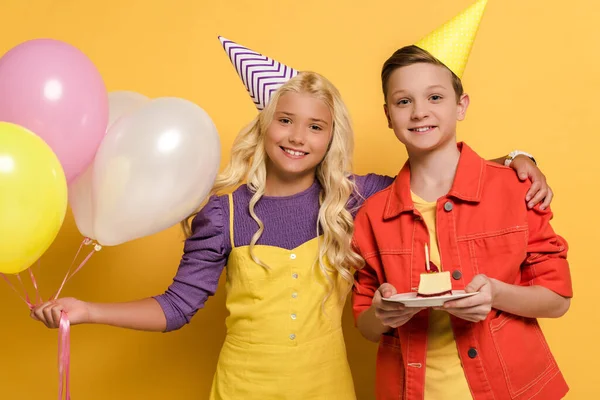 Smiling kids with party caps holding balloons and plate with birthday cake on yellow background — Stock Photo