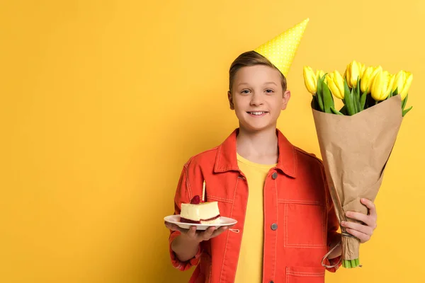 Niño sonriente sosteniendo ramo y plato con pastel de cumpleaños sobre fondo amarillo - foto de stock