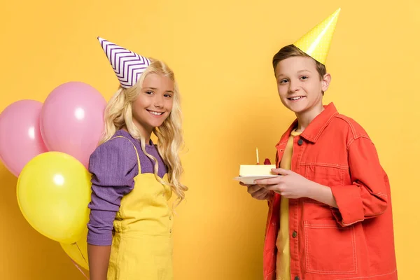 Niños sonrientes con gorras de fiesta sosteniendo globos y plato con pastel de cumpleaños sobre fondo amarillo - foto de stock