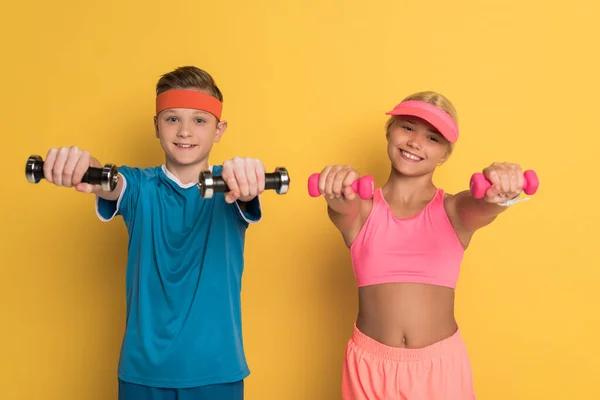 Enfants souriants dans l'entraînement de vêtements de sport avec haltères sur fond jaune — Photo de stock
