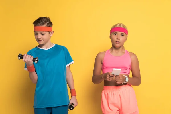 Smiling boy training with dumbbells and his shocked friend holding smartphone on yellow background — Stock Photo