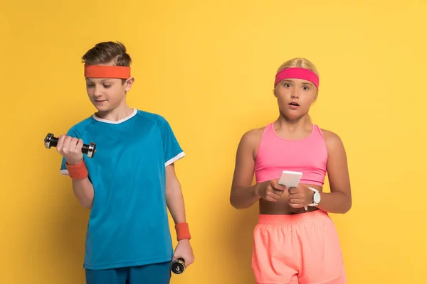 Formation garçon souriant avec haltères et son ami choqué tenant smartphone sur fond jaune — Photo de stock