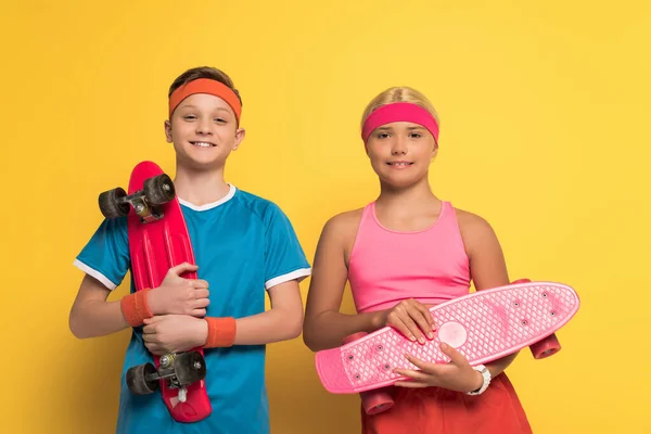 Bambini sorridenti in abbigliamento sportivo tenendo penny board e guardando la fotocamera su sfondo giallo — Stock Photo