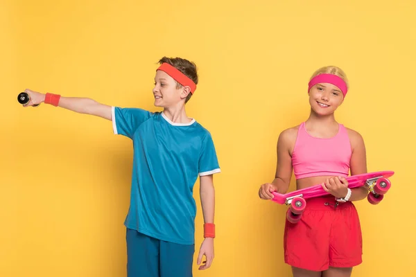 Smiling boy training with dumbbell and his friend  holding penny board on yellow background — Stock Photo