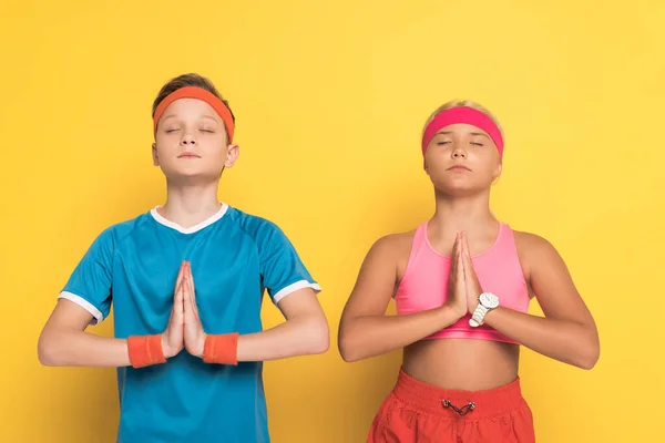 Kids in sportswear with closed eyes showing praying hands on yellow background — Stock Photo
