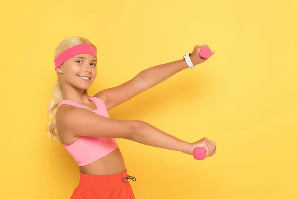 Side view of smiling preteen sportswoman training with dumbbells on yellow background — Stock Photo