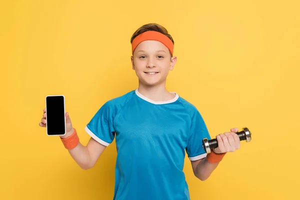 Enfant souriant tenant smartphone et haltère sur fond jaune — Photo de stock