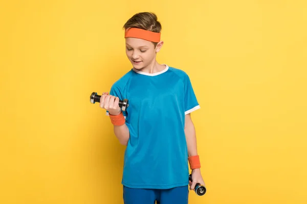 Усміхнена дитина в спортивному одязі тренується з гантелями на жовтому фоні — стокове фото