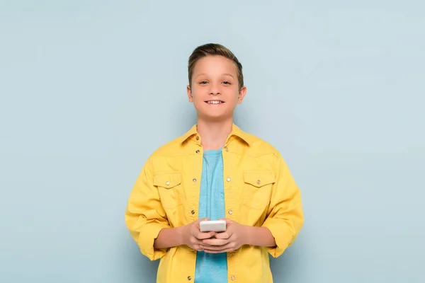 Улыбающийся ребенок, держащий смартфон и смотрящий на камеру на синем фоне — стоковое фото