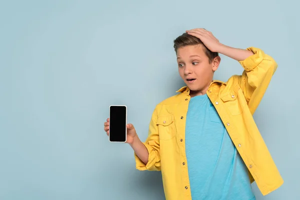 Enfant choqué touchant la tête et tenant smartphone sur fond bleu — Photo de stock