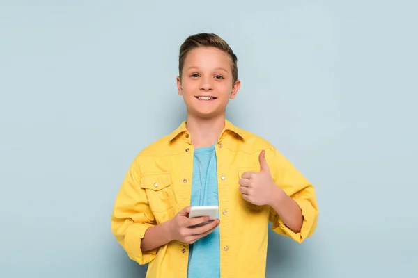 Niño sonriente sosteniendo el teléfono inteligente y mostrando como en fondo azul - foto de stock