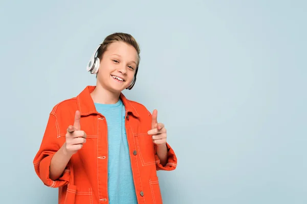 Niño sonriente con auriculares escuchando música y señalando con los dedos sobre fondo azul - foto de stock