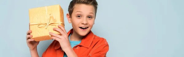 Panoramic shot of smiling kid holding gift box on blue background — Stock Photo