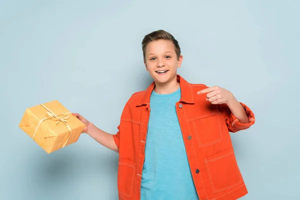 Niño sonriente apuntando con el dedo a la caja de regalo sobre fondo azul - foto de stock