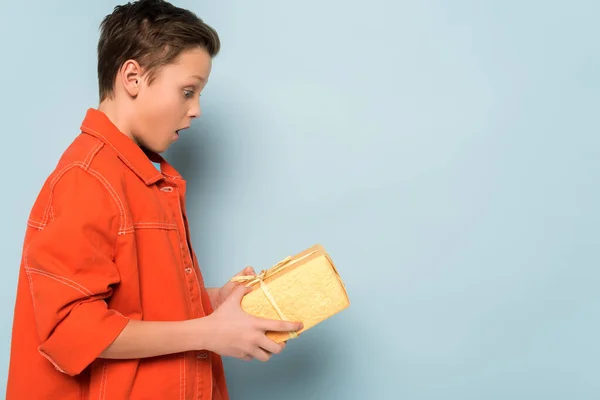 Side view of surprised kid holding gift box on blue background — Stock Photo