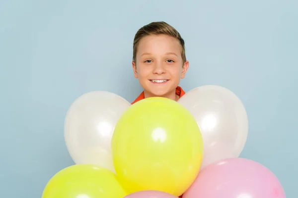 Niño sonriente de pie cerca de globos y mirando a la cámara en el fondo azul - foto de stock