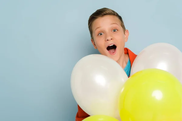 Überraschtes Kind steht in der Nähe von Luftballons und blickt in die Kamera auf blauem Hintergrund — Stockfoto