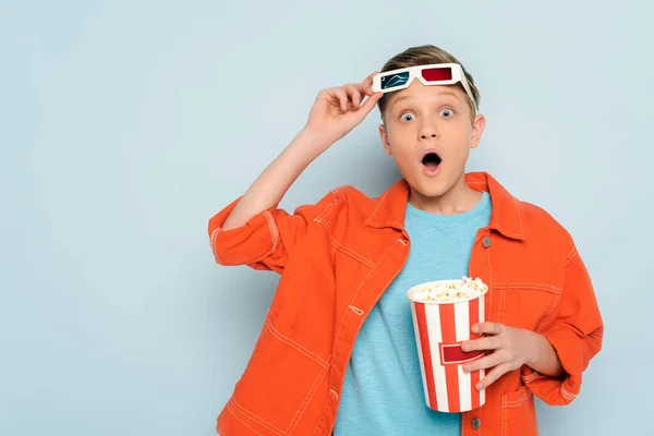 Schockiertes Kind mit 3D-Brille, Eimer mit Popcorn auf blauem Hintergrund — Stockfoto