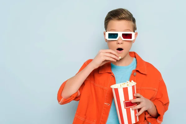 Шокированный ребенок в 3D-очках ест попкорн на синем фоне — стоковое фото