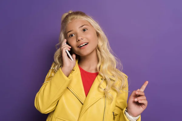 Smiling kid talking on smartphone and showing idea sign on purple background — Stock Photo