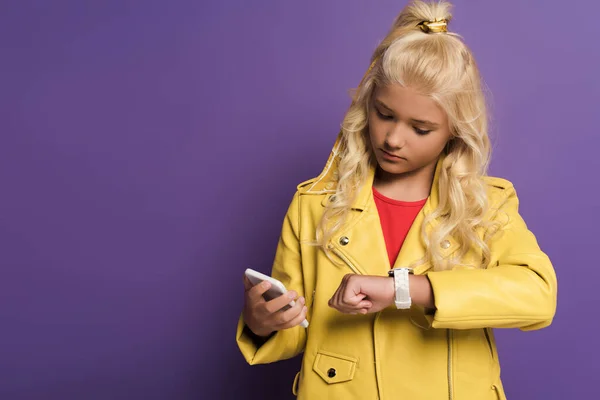 Милый ребенок держит смартфон и смотрит на наручные часы на фиолетовом фоне — стоковое фото