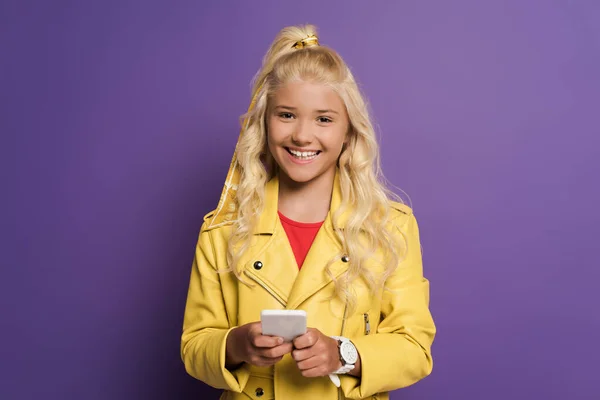 Smiling kid holding smartphone and looking at camera on purple background — Stock Photo