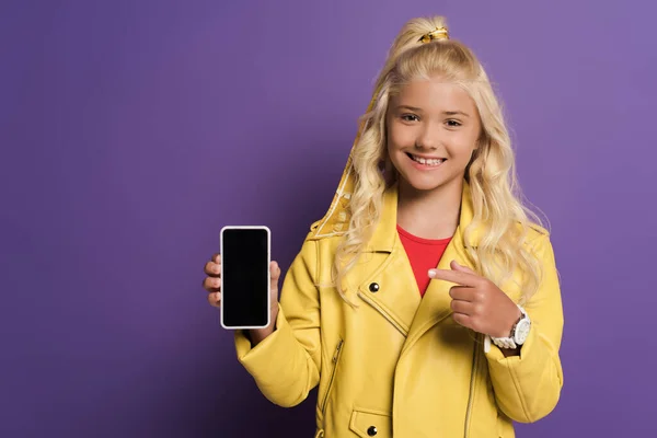 Garoto sorridente apontando com o dedo para o smartphone no fundo roxo — Fotografia de Stock