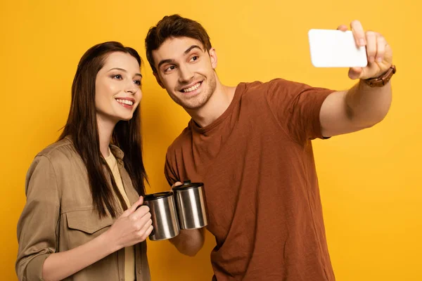 Пара усміхнених мандрівників з чашками з кавою бере селфі на смартфон на жовтому — стокове фото