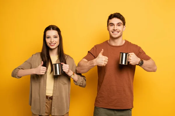 Pareja de viajeros sonrientes sosteniendo tazas con café y mostrando los pulgares hacia arriba en amarillo — Stock Photo