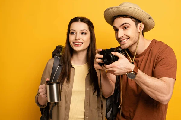 Casal de turistas sorridentes com mochilas segurando câmera fotográfica e café, isolado em amarelo — Fotografia de Stock