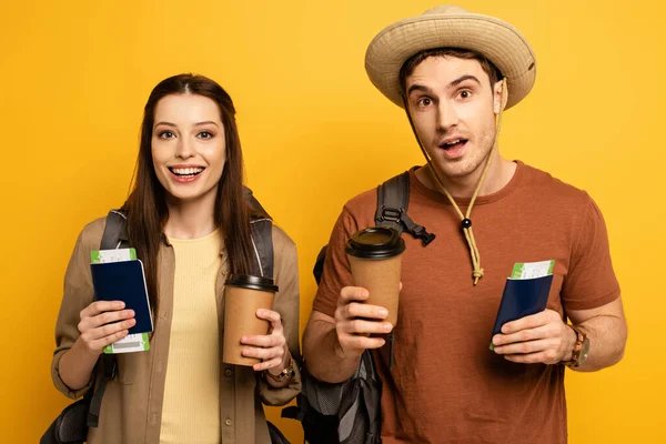 Happy travelers with backpacks holding passports, air tickets and coffee to go on yellow — Stock Photo