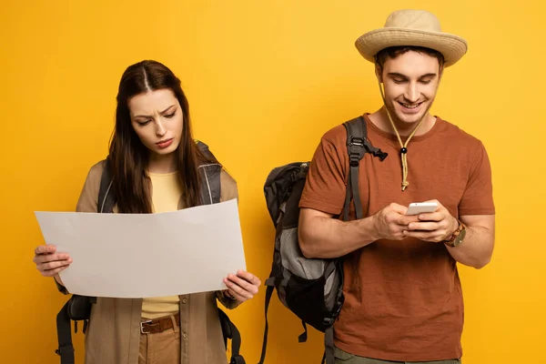 Couple of happy tourists with backpacks looking at map and smartphone on yellow — Stock Photo