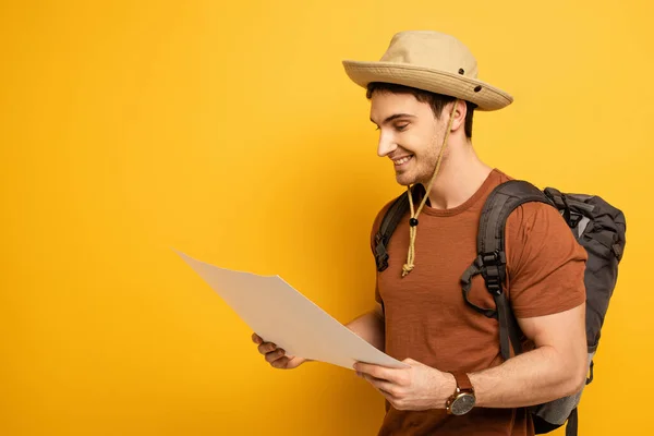 Guapo turista sonriente en sombrero con mochila mirando el mapa en amarillo - foto de stock