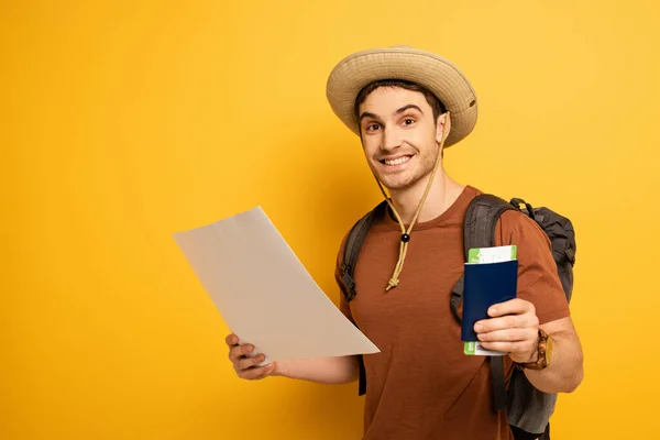 Улыбающийся турист в шляпе с рюкзаком с паспортом, билетом и картой на желтом — стоковое фото