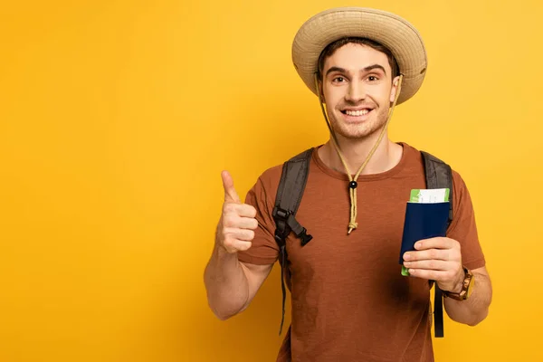 Cheerful tourist in hat with backpack holding passport and ticket while showing thumb up on yellow — Stock Photo