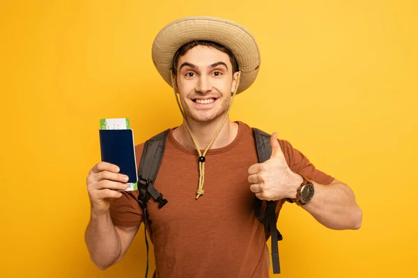 Handsome smiling tourist in hat with backpack holding passport and ticket while showing thumb up on yellow — Stock Photo