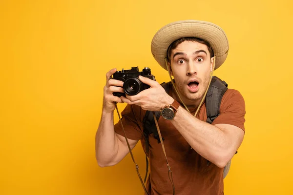 Turista chocado em chapéu com mochila segurando câmera fotográfica no amarelo — Fotografia de Stock