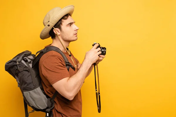 Bellissimo viaggiatore in cappello con zaino in mano fotocamera fotografica su giallo — Foto stock