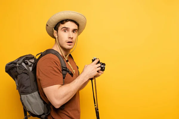 Viajante chocado em chapéu com mochila segurando câmera fotográfica no amarelo — Fotografia de Stock