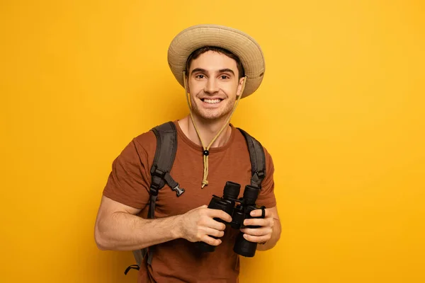 Handsome happy traveler in hat with backpack holding binoculars on yellow — Stock Photo