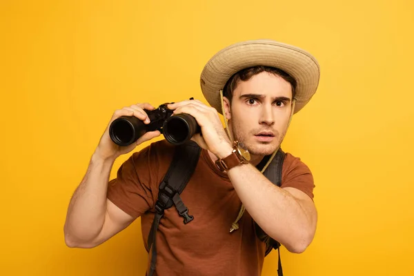 Surprised traveler in hat with backpack holding binoculars on yellow — Stock Photo