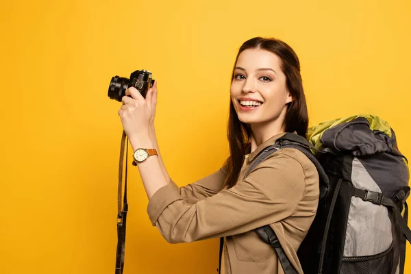 Turista sorridente bonito com mochila segurando câmera fotográfica no amarelo — Fotografia de Stock