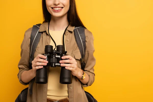 Cropped view of traveler with backpack holding binoculars, isolated on yellow — Stock Photo