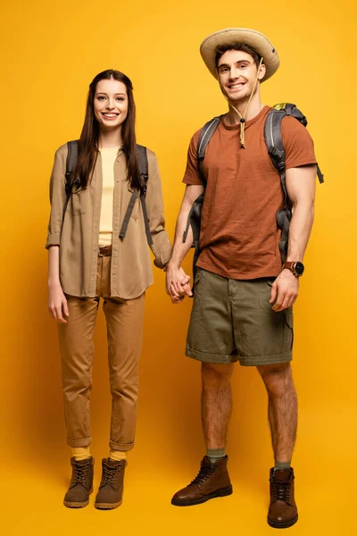 Par de turistas felices con mochilas cogidas de la mano en amarillo - foto de stock