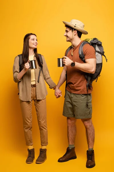Par de turistas felices con mochilas y tazas de café tomados de la mano en amarillo - foto de stock
