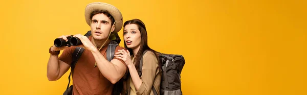 Panoramic shot of  couple of shocked tourists with backpacks and binoculars, Isolated on yellow — Stock Photo