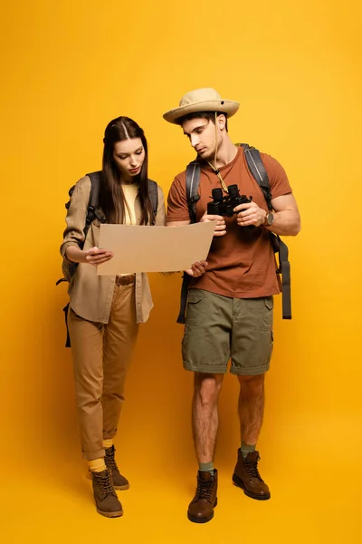 Casal de turistas com mochilas e binóculos olhando para mapa em amarelo — Fotografia de Stock