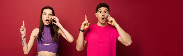 Panoramic shot of shocked couple talking on smartphones while pointing up on red — Stock Photo